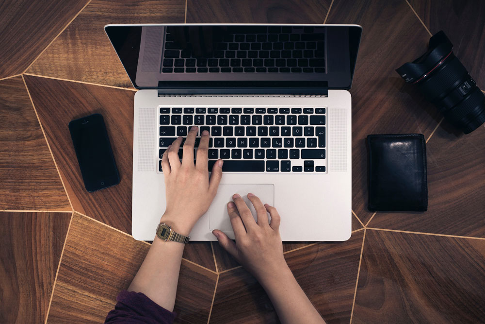 A persons hands on a laptop keyboard with a smartphone and a wallet nearby